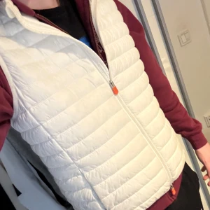 Vit dunväst från Save the Duck - Säljer en stilren vit dunväst från Save the Duck. Västen är quiltad och har en dragkedja framtill. Perfekt för lager-på-lager under kyliga dagar. Den är 100% animal free och ultralätt, vilket gör den både bekväm och miljövänlig. Västen är sparsamt använd och i mycket bra skick! Priset är en hugget i sten!