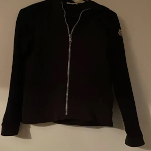 Svart jacka med dragkedja från Moncler - Säljer en svart Moncler jacka med lång ärm och tydlig dragkedja framtill. Klassisk Moncler-logga på ärmen. Enkel och stilren design som passar till många tillfällen.