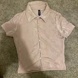 Beige kortärmad skjorta från Divided - Säljer en beige kortärmad skjorta från Divided med klassisk krage och knappar framtill. Skjortan har en lätt glansig yta och är figurnära i modellen. Perfekt för dig som gillar stilrena och enkla plagg.