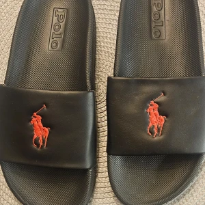 Svarta sandaler från Polo - Snygga svarta sandaler från Polo med röd logotyp av en häst och ryttare på ovandelen. Tillverkade i syntetiskt material med en gummisula för extra komfort. Perfekta för en avslappnad stil.