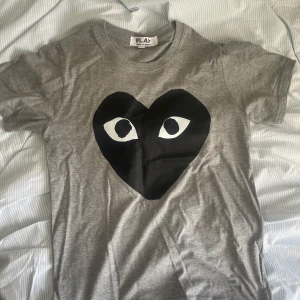 Grå t-shirt från Comme des Garçons - Säljer en grå t-shirt från Comme des Garçons med det ikoniska hjärtat med ögon på framsidan. T-shirten är tillverkad i 100% ull och har en normal passform i storlek S