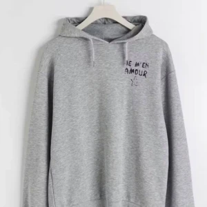 Grå hoodie med text och stjärna - Säljer en stilren grå hoodie med texten 'Je m'en amour' och en stjärna tryckt på både fram- och baksidan. Perfekt för en avslappnad look. Hoodien har en klassisk passform med huva och långa ärmar. Xoxo💕