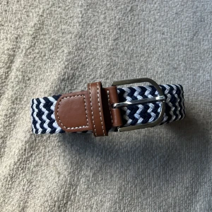 Mörkblå/vit randigt flätat bälte - Snyggt flätat bälte i blått och vitt med bruna läderdetaljer och silverfärgat spänne. Perfekt för att ge en stilren touch till din outfit.