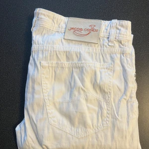 Vita jeans från Jacob Cohen - Säljer ett par vita jeans från Jacob Cohen, handgjorda i Italien med japanska tyger. De har en klassisk design med fem fickor och knappgylf. Perfekta för en stilren look.