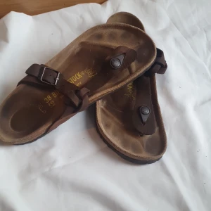 Bruna sandaler från Birkenstock - Säljer ett par klassiska bruna sandaler från Birkenstock med justerbart spänne och tårem. Skorna har en anatomiskt formad fotbädd och robust sula, perfekta för varma dagar.