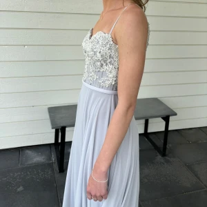 Duvblå långklänning med spets och slits - Säljer en elegant duvblå långklänning med smala axelband, v-ringad baksida med dekorativa knappar. Kjolen är i ett mjukt, flytande tyg med slits. Perfekt för bal eller fest. Använd 2 gånger och har små slitningar längst ut på släpet, men det är inget som syns tydligt. 