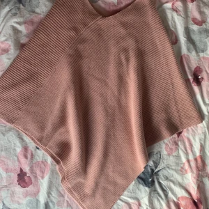 Cape - Mysig ljusrosa stickad poncho från KappAhl. Ponchon har en enkel, klassisk design med ribbade detaljer längs kanterna. Perfekt att slänga över en tröja för extra värme och stil.