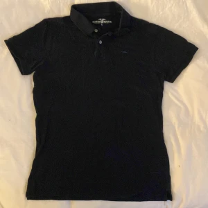 Svart pikétröja från Hampton Republic - Klassisk svart pikétröja från Hampton Republic med krage och två knappar framtill. Tröjan har korta ärmar och en liten broderad logga på bröstet. Perfekt för en stilren och avslappnad look. Nyskick 🫶