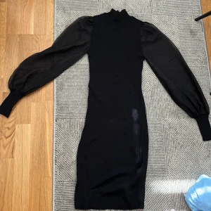 Svart tajt klänning  - Snygg svart klänning med ribbad struktur och puffiga, transparenta ärmar. Den har en hög krage och är figurnära, vilket ger en elegant look. Perfekt för en stilren outfit.