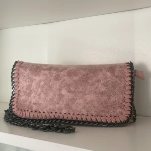 Rosa axelväska med kedja - Säljer en snygg rosa axelväska med grov kedjerem och dekorativ kant i svart. Väskan har ett lock med magnetknappar och dragkedja på insidan. Perfekt för dig som vill ha en stilren accessoar med lite edge.
