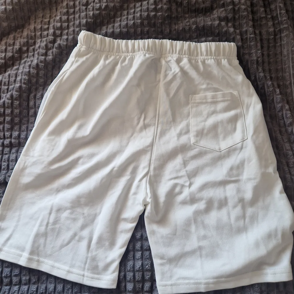 Säljer ett par vita shorts från Essentials Fear of God med svart logga längs benet och snörning i midjan. Perfekta för en avslappnad stil och har elastisk midja för extra komfort. Helt nya och aldrig använda. Storleken är XL men passar som L !. Shortsit.