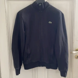 Svart kofta från Lacoste - Säljer en svart kofta från Lacoste med dragkedja och hög krage. Koftan har det klassiska Lacoste-märket broderat på bröstet och två fickor framtill. Perfekt för en stilren och avslappnad look. Säljer då jag växt ur den. 