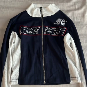 Blå och vit zip-jacka från NVFelix - Säljer en blå och vit crop-top med dragkedja från NVFelix. Crop-toppen har broderad text 'ROCK MORE' och siffran 85 på bröstet. Hög krage och långa ärmar i ribbat material. Perfekt för en cool streetstyle-look. 🎸 Aldrig använd 💙Liten i storleken! (Sitter tajt passar även xs) 