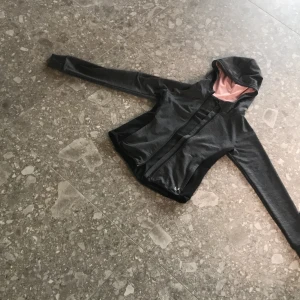 Mörkgrå träningsjacka med huva från Kari Traa - Säljer en mörkgrå träningsjacka med svart logga från Kari Traa. Jackan har huva med rosa foder, dragkedja framtill och långa ärmar. Perfekt för träning utomhus under svalare dagar.