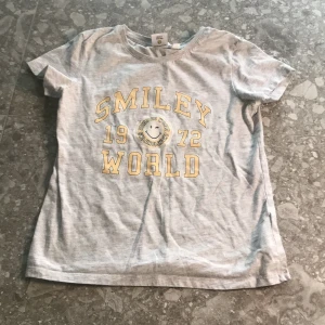 Grå t-shirt från Smiley World - Grå t-shirt från Smiley World med gul text och smiley-tryck på bröstet. Rund halsringning och korta ärmar. Perfekt för dig som gillar en avslappnad och cool stil.