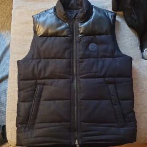 Svart dunväst från Michael Kors - Snygg svart dunväst från Michael Kors med quiltad design och detaljer i skinnimitation på axlarna. Västen har dragkedja framtill, två fickor och diskret MK-logga på bröstet. Perfekt för lager-på-lager under kyliga dagar. 