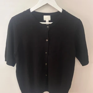 Svart kofta - Säljer en stilren svart kofta från H&M med knappar framtill. Perfekt för lager-på-lager och enkel att matcha med olika outfits. Koftan har korta ärmar och en rund halsringning.