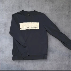 Mörkblå tröja från Hugo Boss - Snygg mörkblå tröja från Hugo Boss med ett stort vitt logotryck på bröstet. Tröjan har långa ärmar och en klassisk rund halsringning. Perfekt för en stilren och avslappnad look.