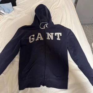Mörkblå hoodie från GANT - Säljer en snygg mörkblå hoodie från GANT med dragkedja och vit logotyp på bröstet. Perfekt för en avslappnad stil. Den har en justerbar huva med snören och långa ärmar.