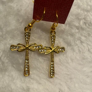 Guldiga korsörhängen med stenar - Snygga örhängen i guldfärg med korsform och glittrande stenar. Ett elegant snirklat band tvärs över korset ger extra detalj. Perfekta för dig som vill sticka ut med accessoarer som syns. Krokarna är i sterling silver 