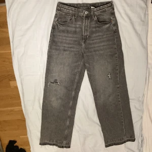 Grå jeans med vida ben från H&M - Säljer ett par grå jeans från H&M med vida ben och hög midja. Byxorna har en justerbar midja och slitna detaljer på framsidan för en trendig look. Perfekta för en avslappnad stil.