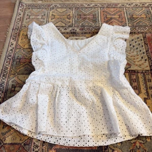 Vit blus med volang från Zara Kids - Söt vit blus från Zara Kids i storlek 152. Blusen har broderade hålmönster, volangdetaljer längs axlarna och en lätt utsvängd nederdel. Perfekt för vår och sommar!