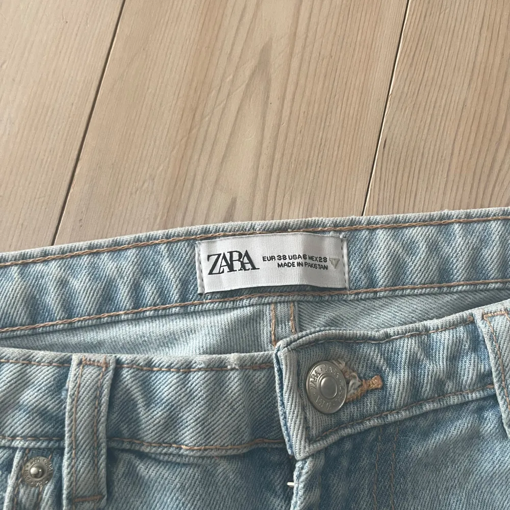 Säljer ett par ljusblå bootcut jeans från Zara med klassisk femficksdesign. Jeansen har en snygg sliten detalj på ena benet och stängs med knapp och dragkedja. Perfekta för dig som gillar en retroinspirerad look.. Farkut & Housut.