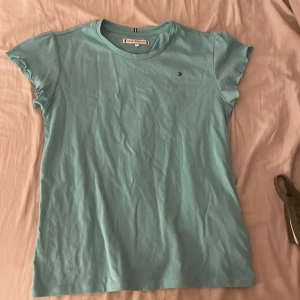 Ljusblå t-shirt från Tommy Hilfiger - Säljer en ljusblå t-shirt från Tommy Hilfiger med korta ärmar och en liten logga på bröstet. Perfekt för en avslappnad stil. Tillverkad i mjukt bomullsmaterial för extra komfort.