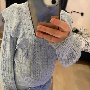 Ljusblå stickad tröja med volangdetaljer - Säljer en ljusblå stickad tröja med vackra volangdetaljer längs ärmarna från Lindex. Tröjan har ett fint stickat mönster och långa ärmar. 