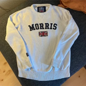 Vit stickad tröja från Morris - Vit stickad tröja från Morris med svart text och brittisk flagga broderad på bröstet. Rund halsringning och ribbade muddar vid ärmslut och nederkant. Perfekt för en stilren och klassisk look.