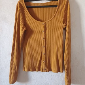 Gul ribbad tröja med knappar - Säljer en snygg gul ribbad tröja med knappar framtill. Tröjan har långa ärmar och en rund halsringning. Perfekt för en stilren look!