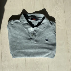 Piké från Morris  - Snygg grå pikétröja med krage och knappar framtill. Den har en diskret broderad detalj på bröstet. Perfekt för en avslappnad stil.