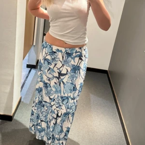 Vit och blå blommig långkjol - Säljer en långkjol med vit bas och blått blommönster. Kjolen har en avslappnad passform och är perfekt för varmare dagar. Den är lätt och luftig, vilket gör den superbekväm att bära.