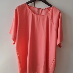 Blus från River Island - Säljer en snygg korallfärgad blus från River Island. Blusen har en lös passform med korta ärmar och en rund halsringning. Den har en liten knappdetalj i nacken för enkel på- och avtagning. Storlek 18. Kan hämtas i Vallentuna.