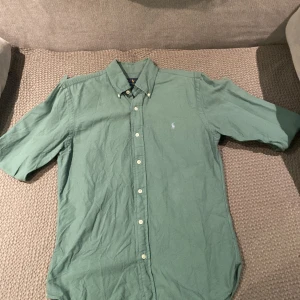 Grön kortärmad skjorta från Ralph Lauren - Säljer en stilren grön kortärmad skjorta från Ralph Lauren i slim fit. Skjortan har en klassisk krage och knappar framtill. Perfekt för en avslappnad men ändå snygg look.