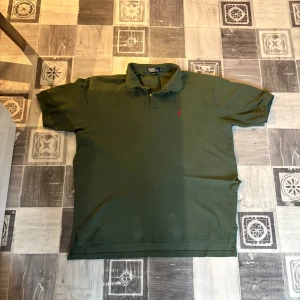 Olivgrön pikétröja från Polo Ralph Lauren - Säljer en olivgrön pikétröja från Polo Ralph Lauren i storlek XL. Tröjan är tillverkad i 100% bomull och har korta ärmar samt klassisk krage med knappar. Liten broderad logga på bröstet. Perfekt för en avslappnad och stilren look.