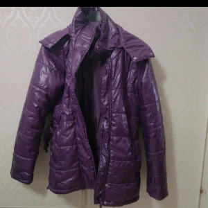 Lila quiltad pufferjacka med bälte - Säljer en lila quiltad pufferjacka med avtagbart bälte och huva. Jackan har hög krage, långa ärmar och stängs med dragkedja och knappar. Perfekt för kyligare dagar och ger en snygg siluett.