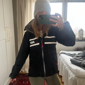 Svart jacka från Helly Hansen - Säljer en svart jacka från Helly Hansen med vit logga och detaljer. Jackan har en röd huva och dragkedja framtill. Perfekt för kyliga dagar med sin långa ärmar och funktionella design.