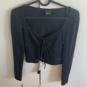 Svart långärmad topp från Gina Tricot - Snygg svart långärmad topp från Gina Tricot med knytning framtill. Perfekt för en stilren look. Toppen har en v-ringad design och är tillverkad i ett mjukt material.
