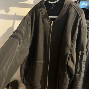 Svart bomberjacka med dragkedja - Svart bomberjacka från H&M med klassisk passform. Jackan har ribbade muddar, dragkedja framtill och fickor på sidorna. Ärmarna är i glansigt material med en extra ficka och dekorativa detaljer. 