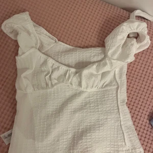 Vit blus med volang från Primark - Supersöt vit blus från Primark med volangdetaljer vid axelbanden och lätt rynkad byst. Toppen har ett diskret rutmönster i tyget och breda axelband. Perfekt för dig som gillar   luftig stil. Storlek XS.