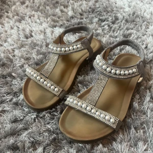 Beige sandaler med pärlor och strass - Snygga beige sandaler med elastisk hälrem, dekorerade med stora pärlor och glittrande strass på remmarna. Platt sula och öppen tå, de är i storlek 38 och jag fick dom i födelsedags present så vet ej vart dom är ifrån 💕väldigt fina när man tex är utomlands eller bara att ha på en kväll 💕säljer då de är för små för mig. De är lite ställen där de är lite slottet men de syns på bilden💕