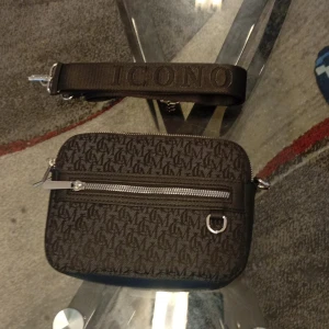 Brun axelväska från Icono med monogram - Snygg brun axelväska från Icono med präglat monogrammönster över hela väskan. Väskan har en bred axelrem med broderad logga, silverfärgade dragkedjor och detaljer samt en praktisk ytterficka framtill. Perfekt storlek för mobilen och andra essentials.