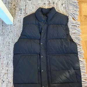 Svart dunväst med stora fickor - En svart dunväst med hög krage och stora, snedställda fickor framtill. Västen har tryckknappar och dragkedja, quiltad design och en clean look utan synliga loggor. Perfekt för lager-på-lager under kyliga dagar.