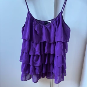 Lila volanglinne från H&M Divided - Supersött lila linne från H&M Divided med tunna axelband och flera lager volanger som ger en riktigt flowy och lekfull look. Perfekt för dig som vill sticka ut med färg och volym. Luftigt material som känns lätt på kroppen.