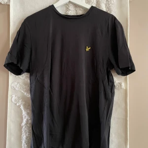 Svart t-shirt från Lyle & Scott - Svart t-shirt från Lyle & Scott med klassisk rund hals och korta ärmar. På bröstet sitter den ikoniska gula fågeln broderad. Tillverkad i mjuk bomull som känns skön mot huden. Perfekt för en clean och stilren look.