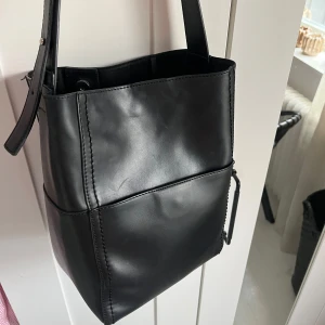 Svart axelväska i skinn - Snygg svart axelväska i slätt skinn med stilren, fyrkantig form från Åhléns. Väskan har bred axelrem, magnetlås och flera innerfack med dragkedja för smidig organisering. Perfekt för dig som vill ha en clean och klassisk look. Använda en del men är fortfarande i okej skick. Skicka för fler bilder eller frågor🩷