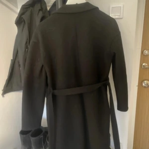 Svart kappa från Pieces med bälte - Svart lång kappa från Pieces med klassisk krage och bälte i midjan. Kappan har raka ärmar, stora fickor framtill och en stilren look som passar till många outfits. Perfekt för kyligare dagar och enkel att styla.