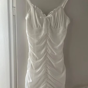 Vit kortklänning - Supersöt vit kortklänning med smala axelband och rynkade detaljer framtill och baktill. Klänningen har även en liten rosett vid urringningen. Perfekt för dig som vill sticka ut med en stilren look.