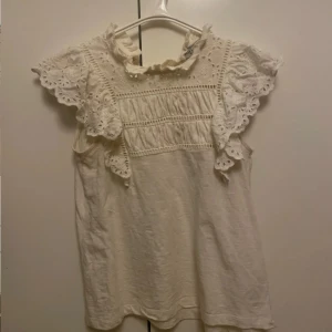 Vit blus med spets från Holly & Whyte - Supersöt vit blus från Holly & Whyte med volangiga ärmar och broderade spetsdetaljer. Hög krage med liten volang och fina hålbroderier på axlar och ärmar. Perfekt för dig som gillar romantisk och feminin stil.